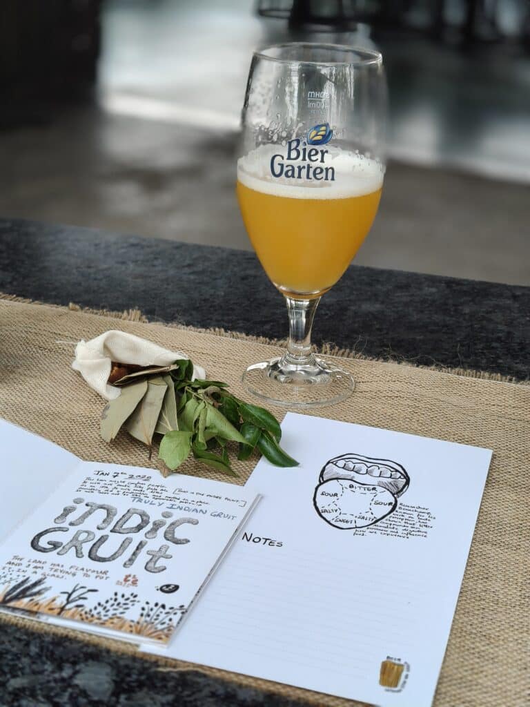 Half-full glass of Biergarten Brewery's Indic Gruit with printed tasting notes and small quantities of bay leaf and lemon leaf in the foreground.
